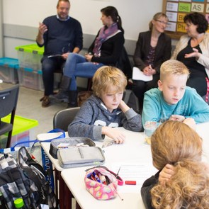 Kinder und Lehrer sitzen locker verstreut im Klassenraum und sprechen miteinander