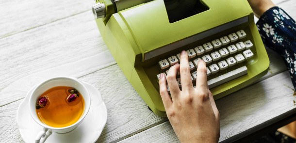 Eine Frau spannt ein Blatt Papier in eine Schreibmaschine ein, neben der Schreibmaschine steht eine Tasse mit Tee auf dem Tisch.