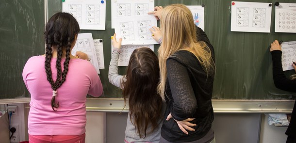 Eine Lehrerin und zwie ca. 7 und 10 jährige Kinder kleben etwas an eine Schultafel