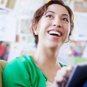 Eine junge Frau sitzt mit einem Tablet in der Hand auf dem Sofa und lacht fröhlich eine nicht im Bild sichtbare Person an.