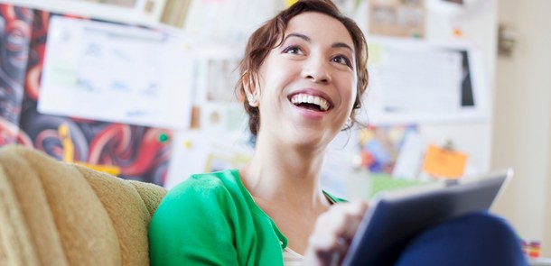 A young woman sits on a sofa, holding a tablet PC and smiling happily towards a person who is not visible in the picture.