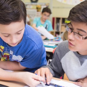 Zwei Jungs sitzen nebeneinander, ein Junge erklärt dem anderen etwas, sie schauen in ein Schulheft