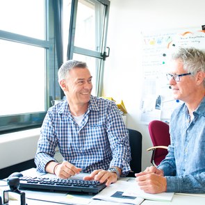zwei Männer sitzen an einem Schreibtisch nebeneinander und sprechen miteinander