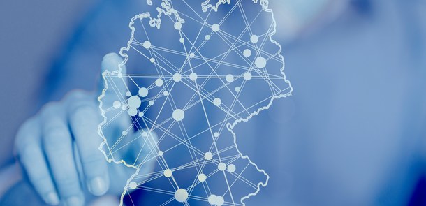 Eine blaue Glaswand. Auf der Glaswand ist Deutschland als Kartengrafik dargestell. Städte sind mit weißen Punkten markiert, die untereinander mit weißen Linien verbunden sind. Hinter der Glaswand steht jemand und zeigt mit dem Finger auf die Kartengraf