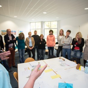 Seminarteilnehmer stehen in einem Kreis um einen Gruppentisch, während der Seminarleiter Erklärungen gibt.